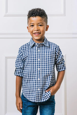 A smiling child models a navy gingham button-down shirt with rolled sleeves, paired with blue jeans against a white background.