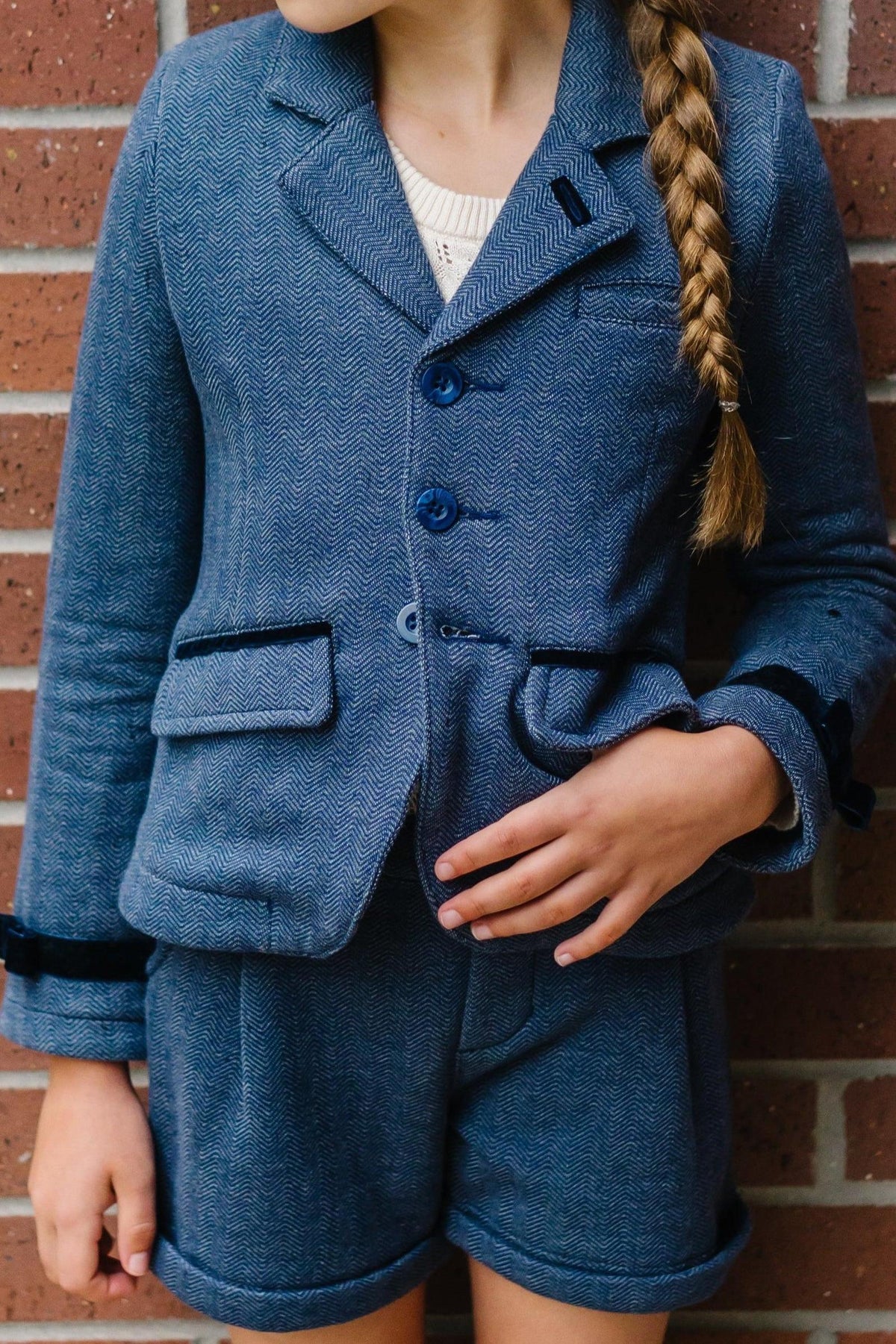 A young girl wears a stylish blue herringbone blazer with matching shorts, standing against a brick wall background.
