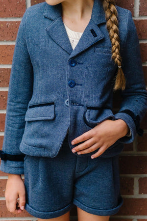 A young girl wears a stylish blue herringbone blazer with matching shorts, standing against a brick wall background.