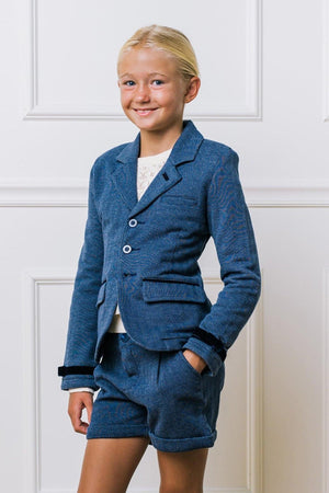 A young girl smiles while wearing a blue herringbone blazer and matching shorts, set against a light-colored wall.