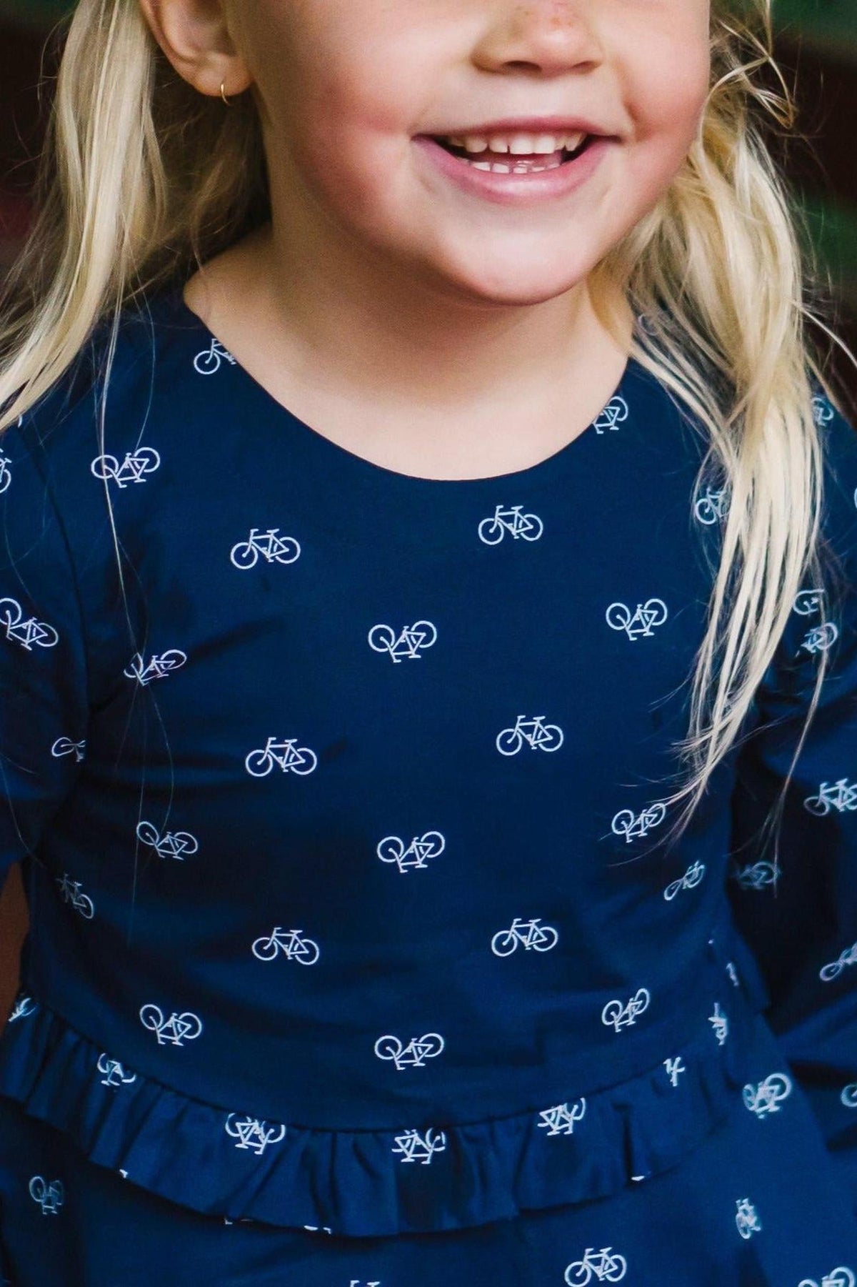 A smiling child wearing a navy dress with a bicycle print, featuring a ruffle detail at the waist.