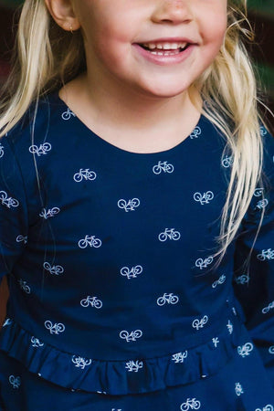 A smiling child wearing a navy dress with a bicycle print, featuring a ruffle detail at the waist.