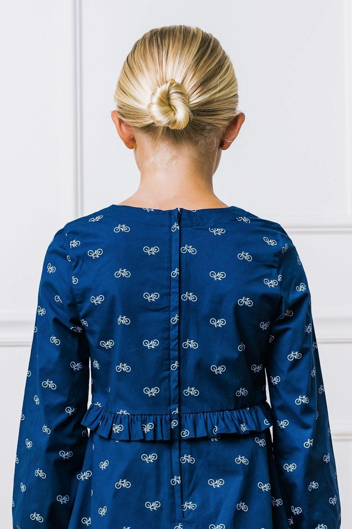 A young girl’s back is shown, wearing a navy dress with a bicycle print and a ruffled waist detail.