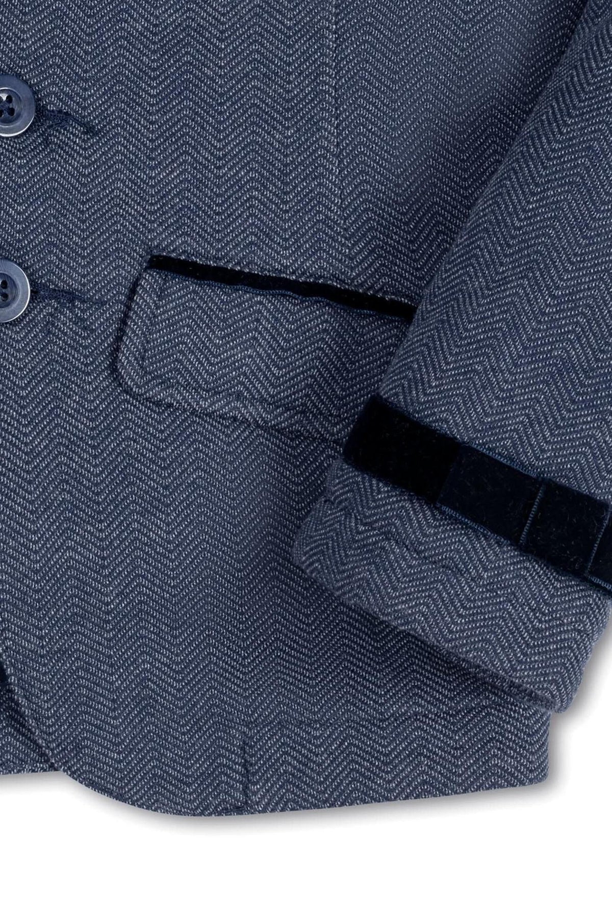 Close-up of a navy blue blazer featuring a herringbone pattern, faux suede piping, and decorative bow at the cuff.