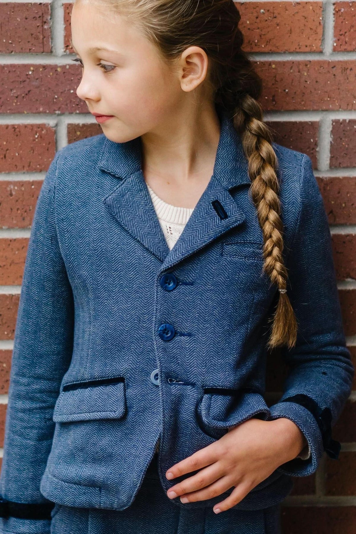 A girl in a blue herringbone blazer with faux suede accents stands against a brick wall, showcasing a stylish layered.