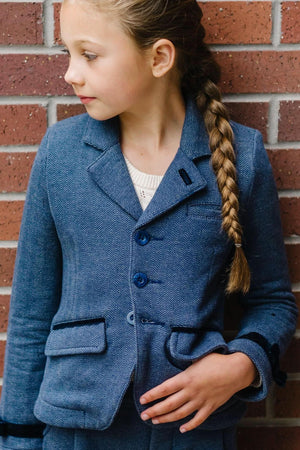 A girl in a blue herringbone blazer with faux suede accents stands against a brick wall, showcasing a stylish layered.