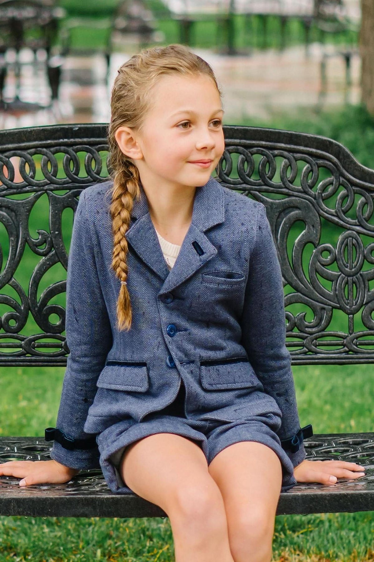 A young girl sits on a decorative bench, wearing a blue herringbone blazer and a light top, smiling softly.
