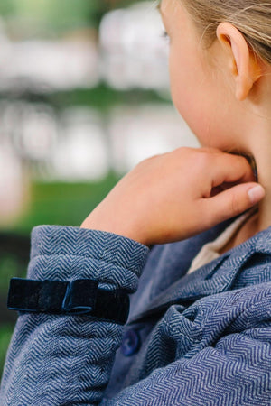 A close-up of a childs arm wearing a textured blue blazer with a dark velvet bow on the cuff.