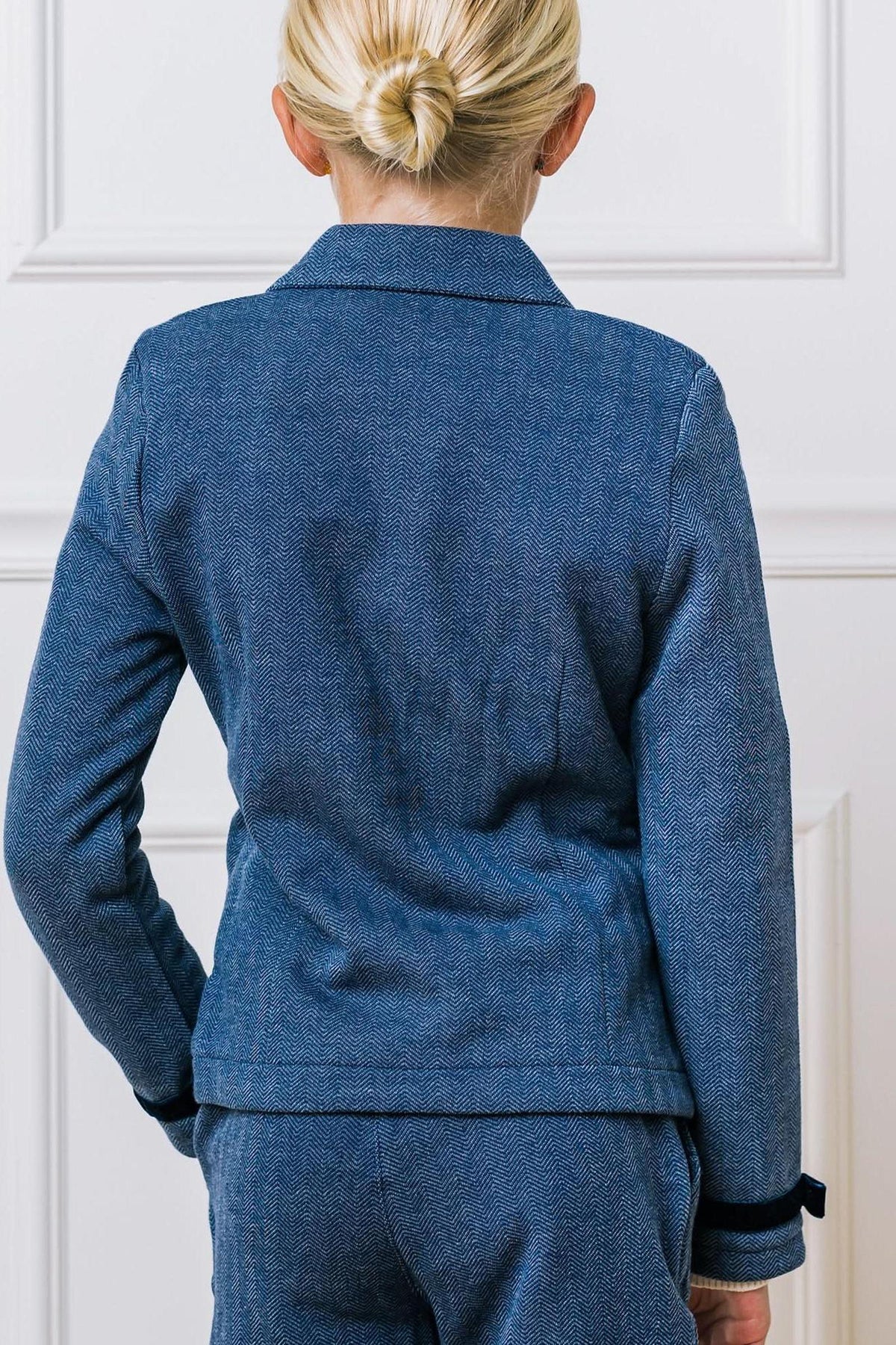 A young girl is seen from the back, wearing a blue herringbone-patterned blazer with long sleeves and a classic collar.