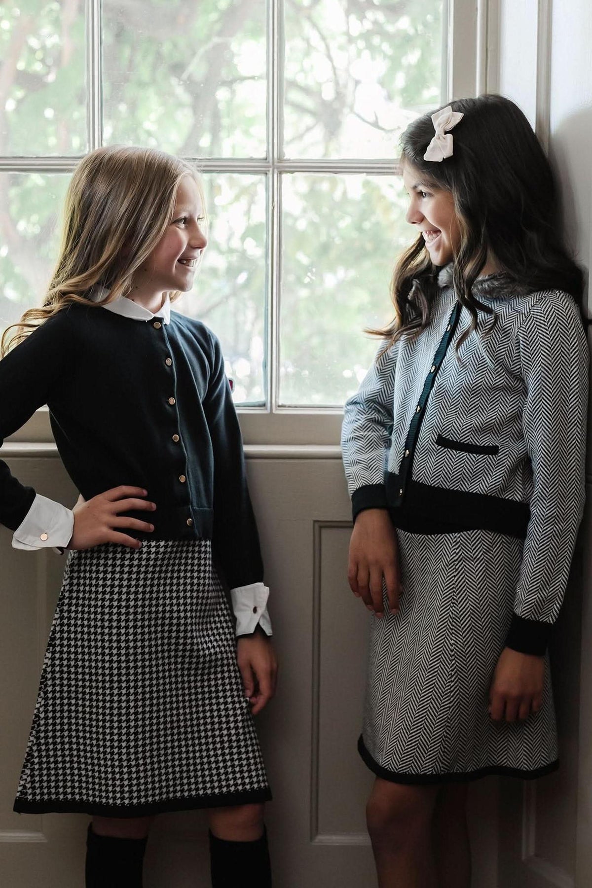 Two girls smiling by a window, wearing stylish herringbone and houndstooth sweater sets in a well-lit room.