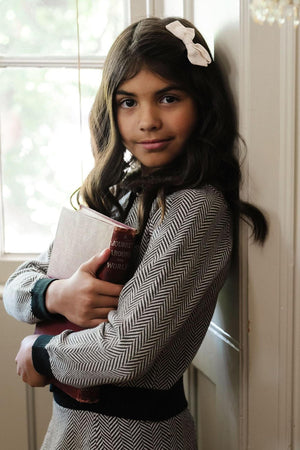 A girl wearing a herringbone-patterned sweater holds a book, standing by a window with soft natural light.