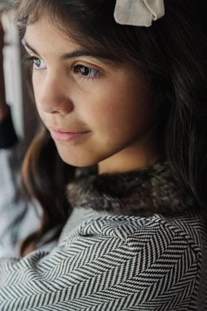 A girl with long dark hair wears a herringbone-patterned cardigan with a faux fur collar and a white bow.