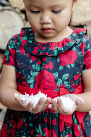 A young girl in a black and red floral dress holds white snow in her hands, displaying a thoughtful expression.