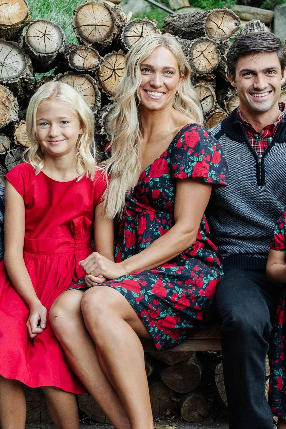 A woman in a floral dress sits beside a girl wearing a red dress, both smiling in front of a stacked log backdrop. The scene captures a warm, family-friendly moment.