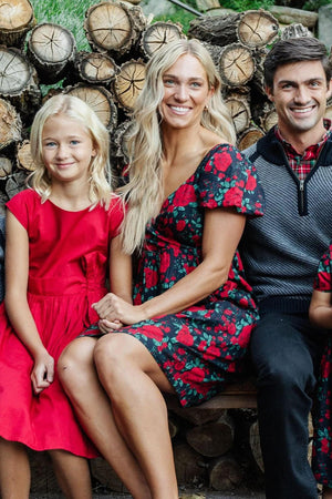 A woman in a floral dress sits beside a girl wearing a red dress, both smiling in front of a stacked log backdrop. The scene captures a warm, family-friendly moment.