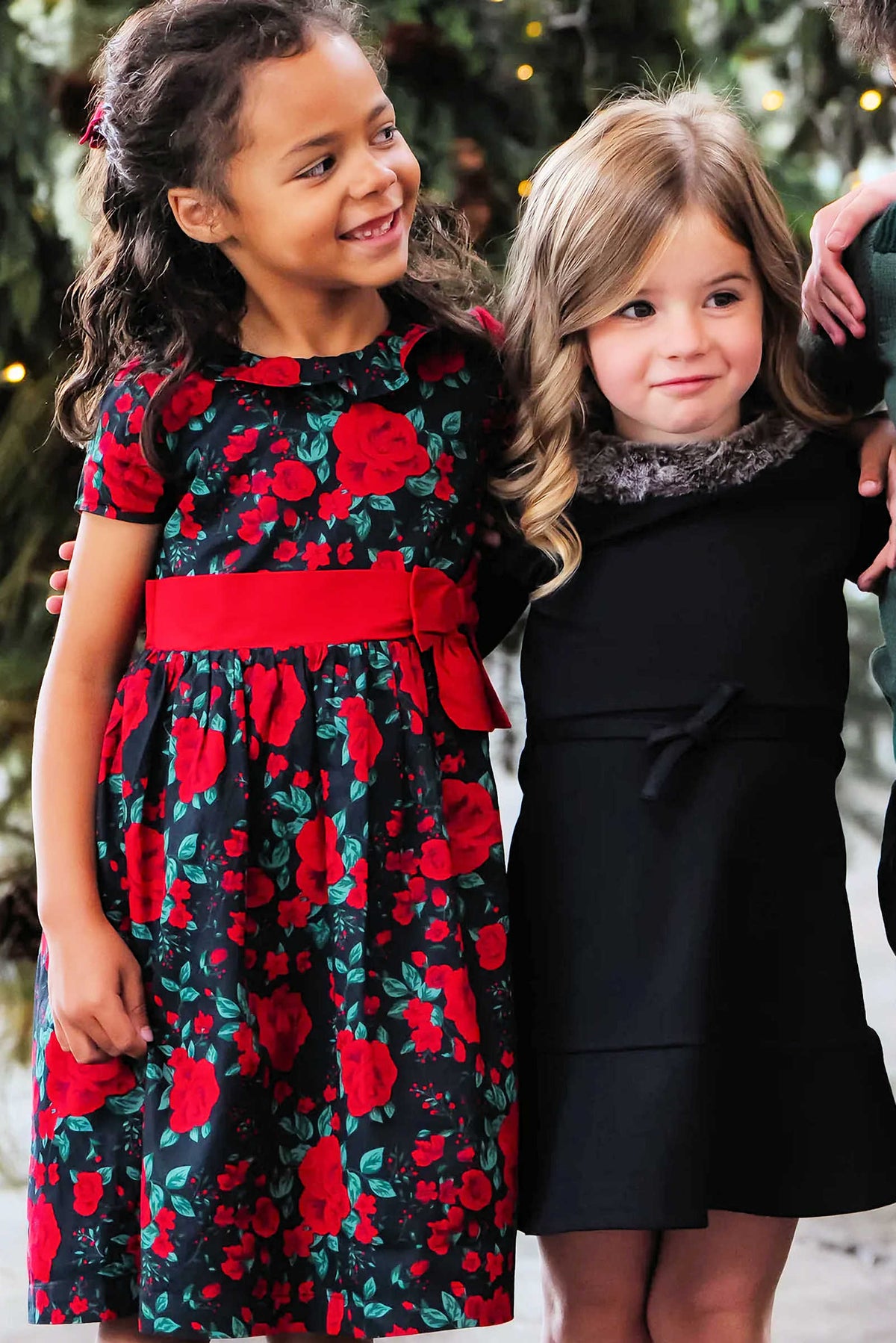 Two young girls stand together, one wearing a black and red floral dress with a waist sash, while the other dons a simple black dress with a stylish collar.