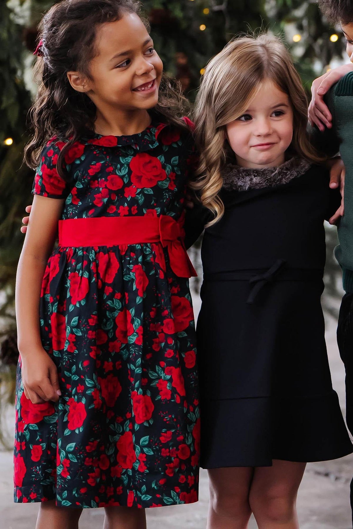 Two young girls stand together in festive attire. One wears a black dress adorned with red roses and a red waist sash, while the other sports a simple black dress.
