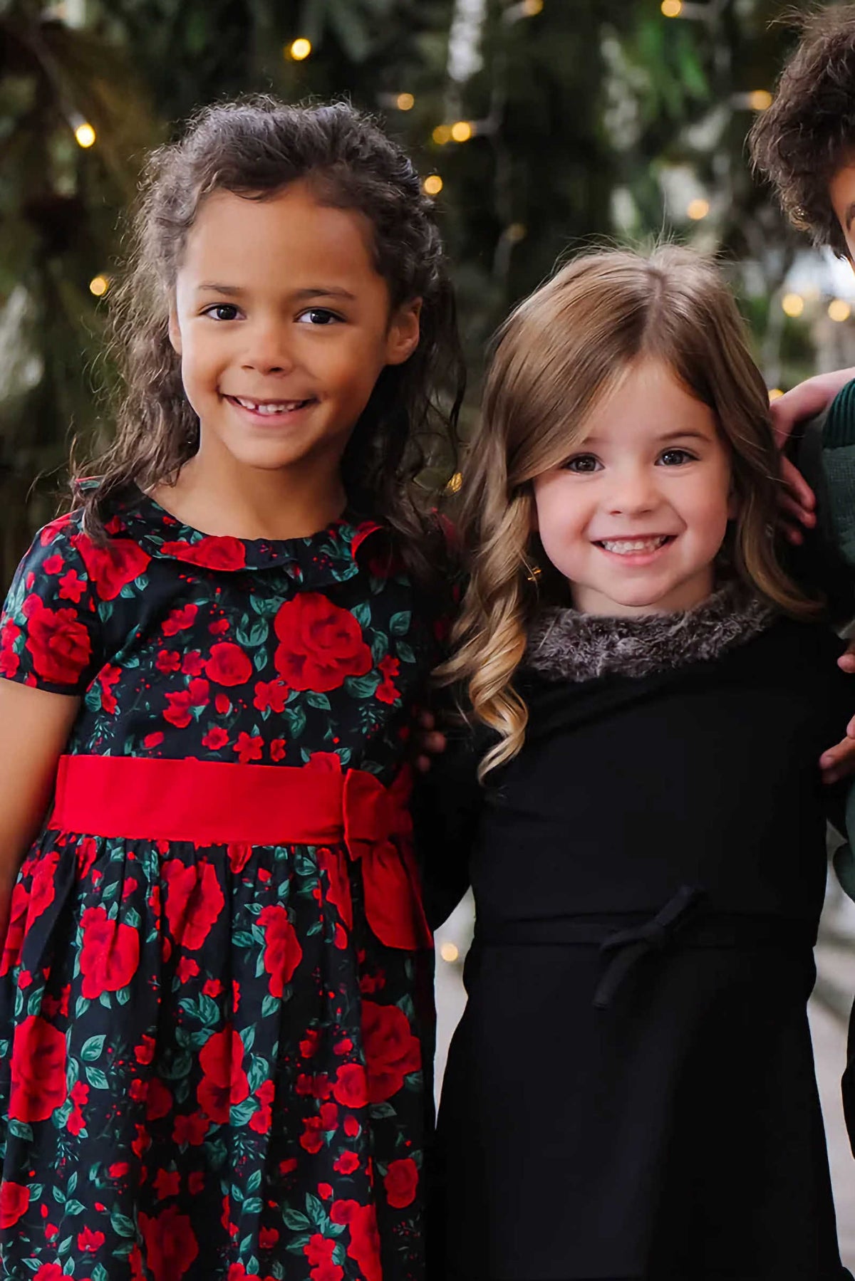 Two young girls smile together, one wearing a black dress with a ruffle collar and the other in a floral red and black dress with a red waist sash.