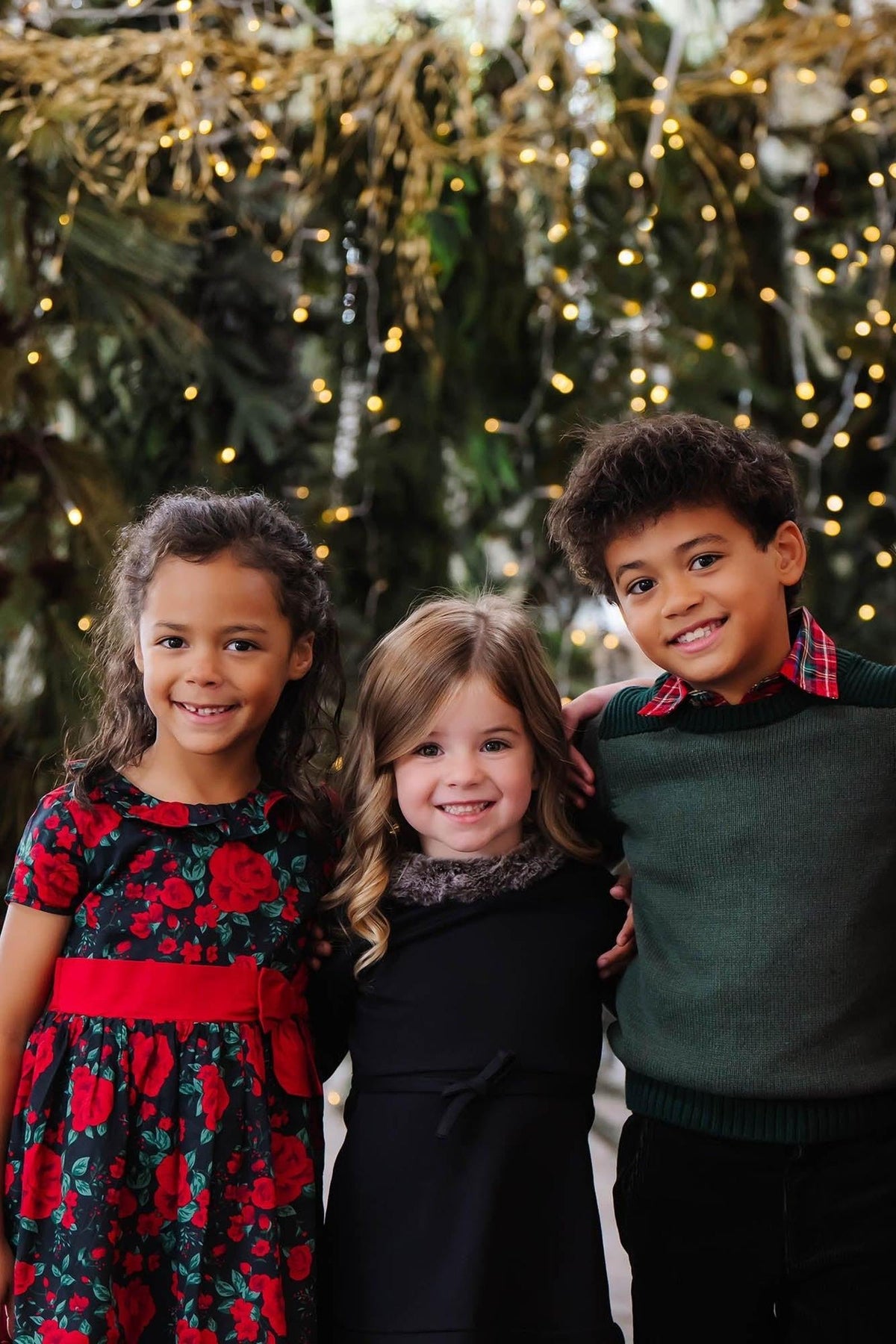 Three smiling children stand together, surrounded by festive greenery and twinkling lights. They wear colorful holiday outfits, including floral patterns and cozy knits, perfect for joyful celebrations.