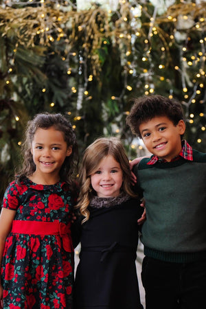 Three smiling children stand together, surrounded by festive greenery and twinkling lights. They wear colorful holiday outfits, including floral patterns and cozy knits, perfect for joyful celebrations.