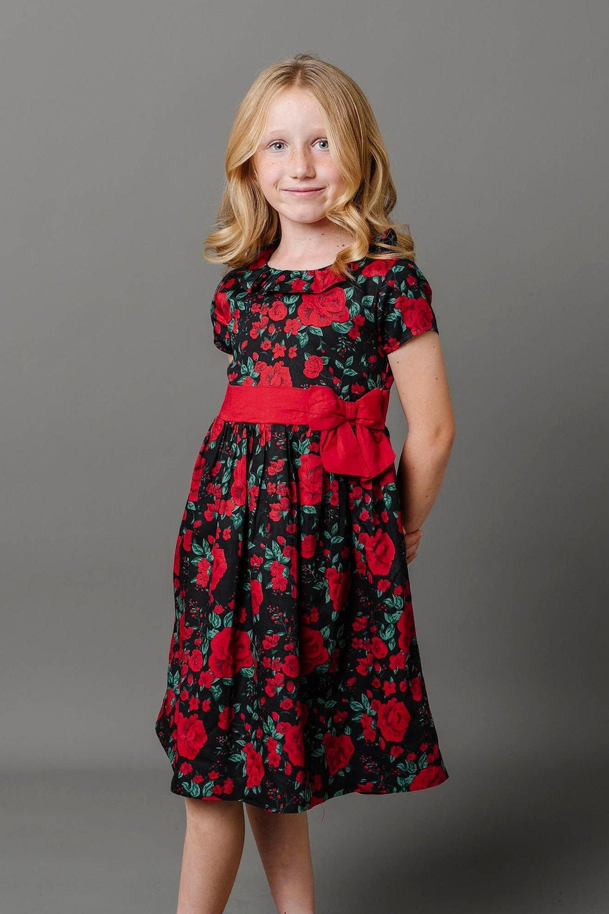 A young girl poses in a black floral party dress featuring red roses, a ruffle collar, and a waist sash with a bow.