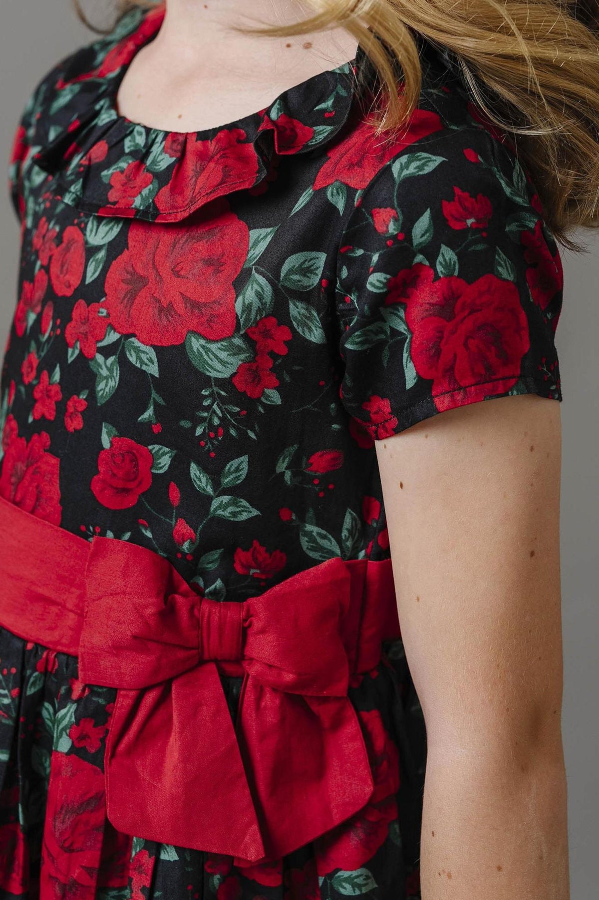A close-up view of a girls black and red floral dress featuring a ruffle collar and a red sash with a bow.