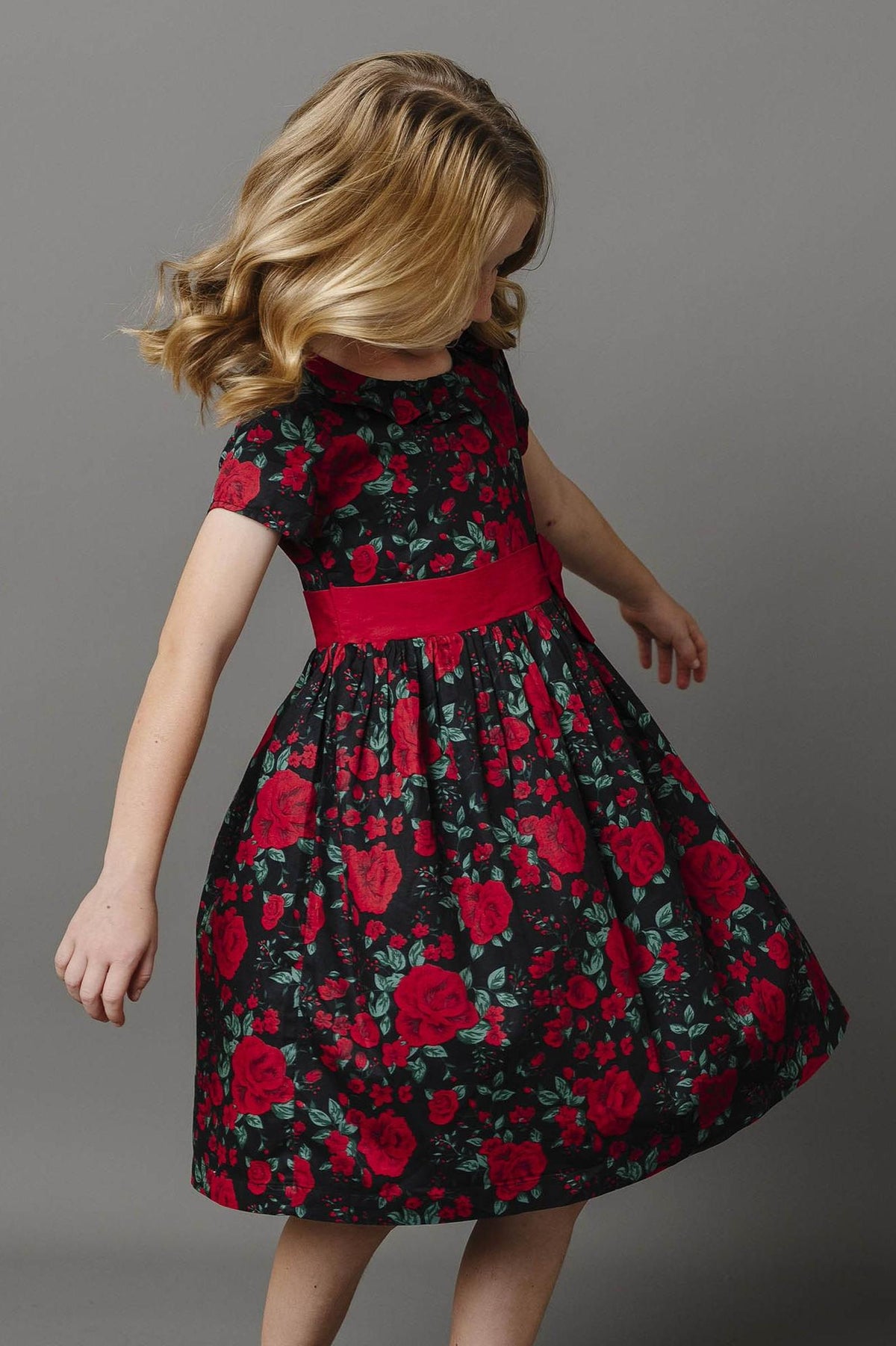 A young girl twirls in a black dress adorned with vibrant red roses, featuring a ruffle collar and a matching waist sash. The dress is made from organic cotton.