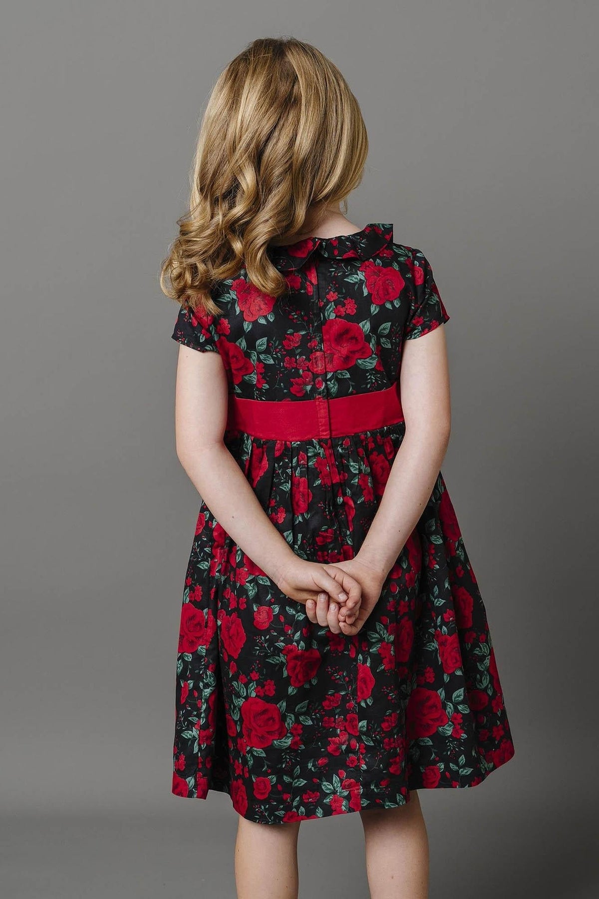 A young girl stands with her back to the camera, wearing a black dress adorned with red floral patterns and a red waist sash.