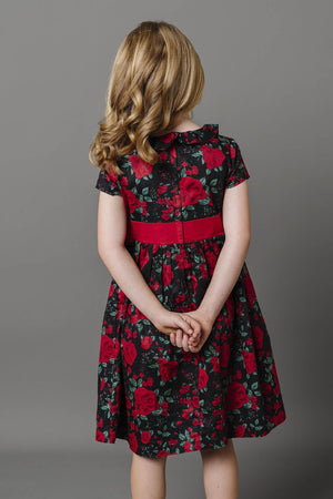 A young girl stands with her back to the camera, wearing a black dress adorned with red floral patterns and a red waist sash.