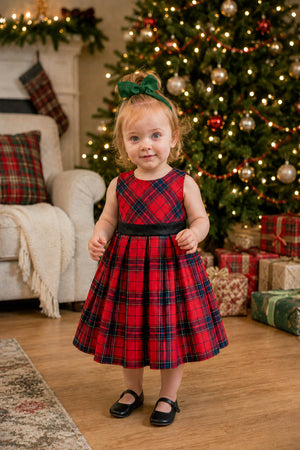 A toddler girl stands in a festive setting, wearing a red holiday plaid pleated dress with a black waistband.