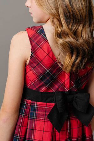 A girl models a red tartan pleated dress featuring a fitted bodice and a black waist sash with a bow.