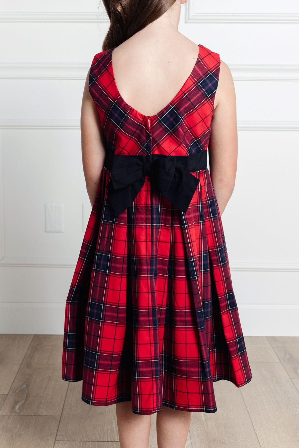 A young girl wears a red tartan pleated dress with a fitted bodice and a black bow at the waist, showcasing the elegant back design against a neutral background.