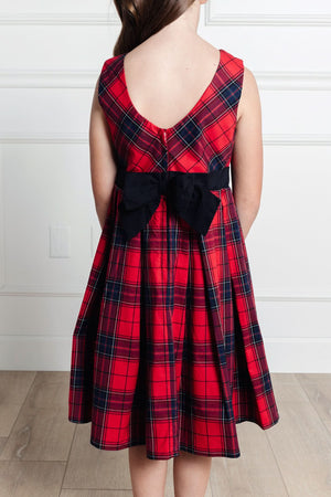 A young girl wears a red tartan pleated dress with a fitted bodice and a black bow at the waist, showcasing the elegant back design against a neutral background.
