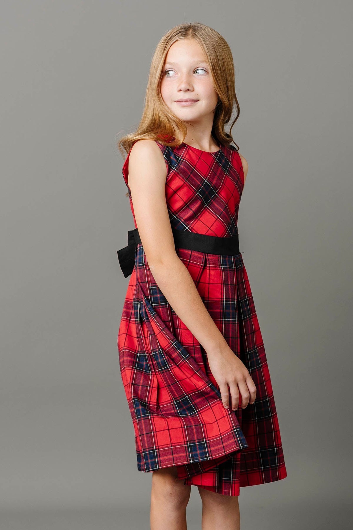 A young girl poses in a vibrant red tartan dress featuring a fitted bodice, A-line skirt, and a black waist sash with a bow, set against a neutral background.