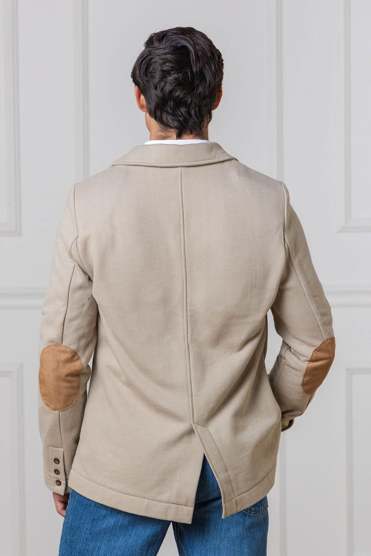 A man stands with his back to the camera, wearing a taupe cotton fleece blazer featuring faux suede elbow patches and a back vent, set against a neutral, indoor backdrop.