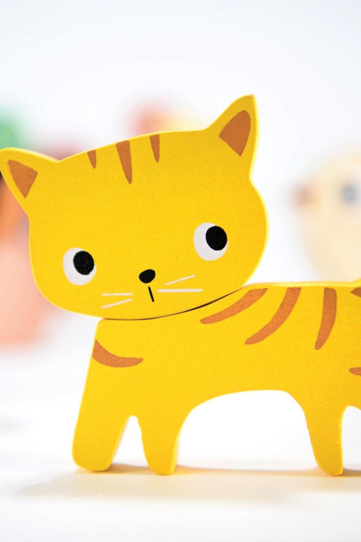 Yellow wooden cat toy with simple painted features, set against a blurred background. Suitable for toddlers.