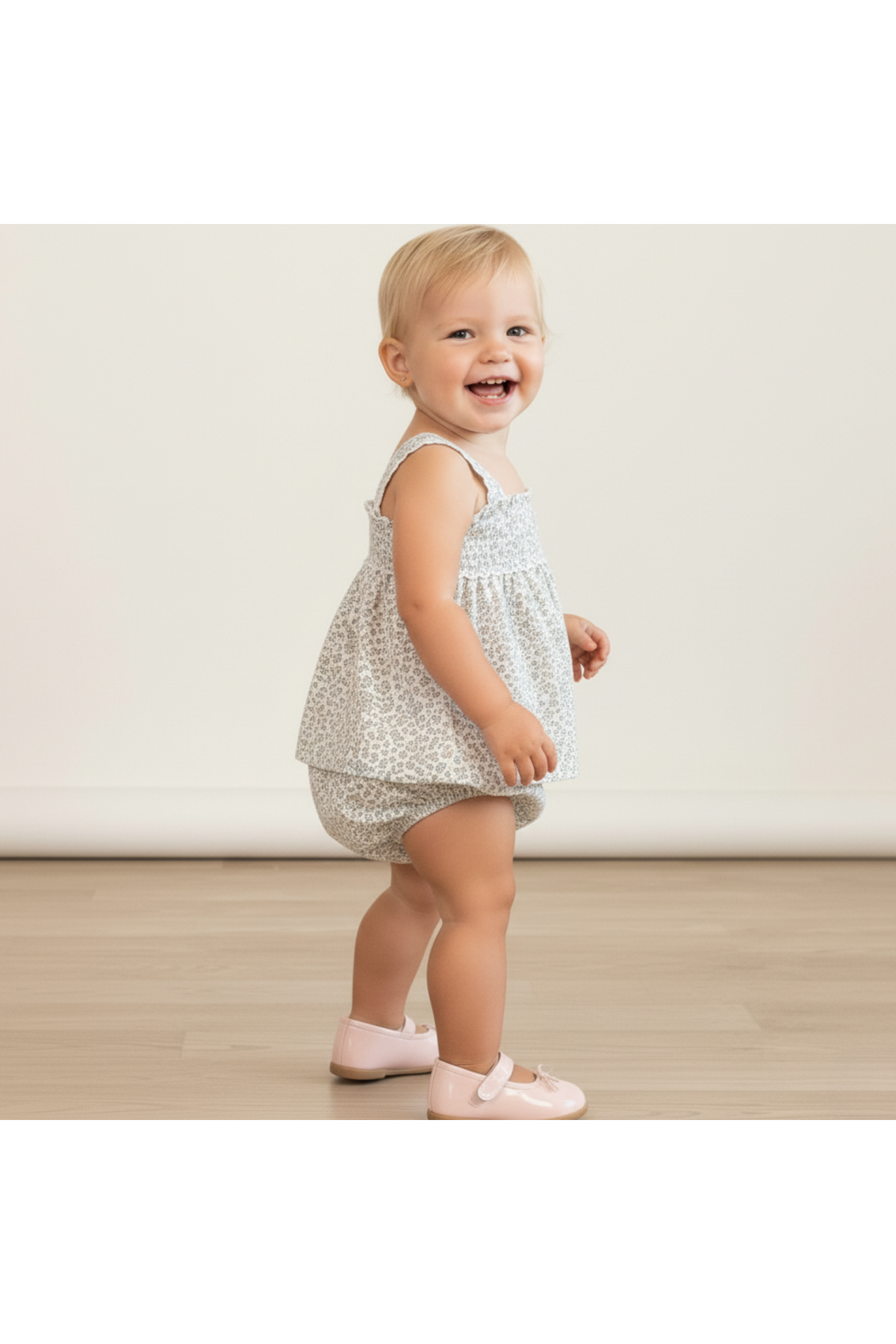 A smiling toddler stands sideways, wearing a light floral-patterned top and matching bloomers. The outfit features delicate straps and a relaxed fit, perfect for warm weather and playful moments.