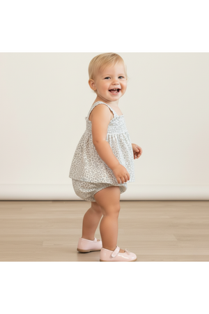 A smiling toddler stands sideways, wearing a light floral-patterned top and matching bloomers. The outfit features delicate straps and a relaxed fit, perfect for warm weather and playful moments.