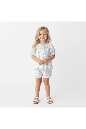 A young girl stands wearing a blue and white organic cotton smocked top and shorts set, featuring delicate floral patterns. The outfit is playful and perfect for sunny day outings.