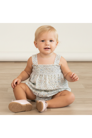A smiling toddler sits on a light wooden floor, wearing a floral-patterned romper with adjustable straps. The outfit features soft, breathable fabric, perfect for warm weather and active play.
