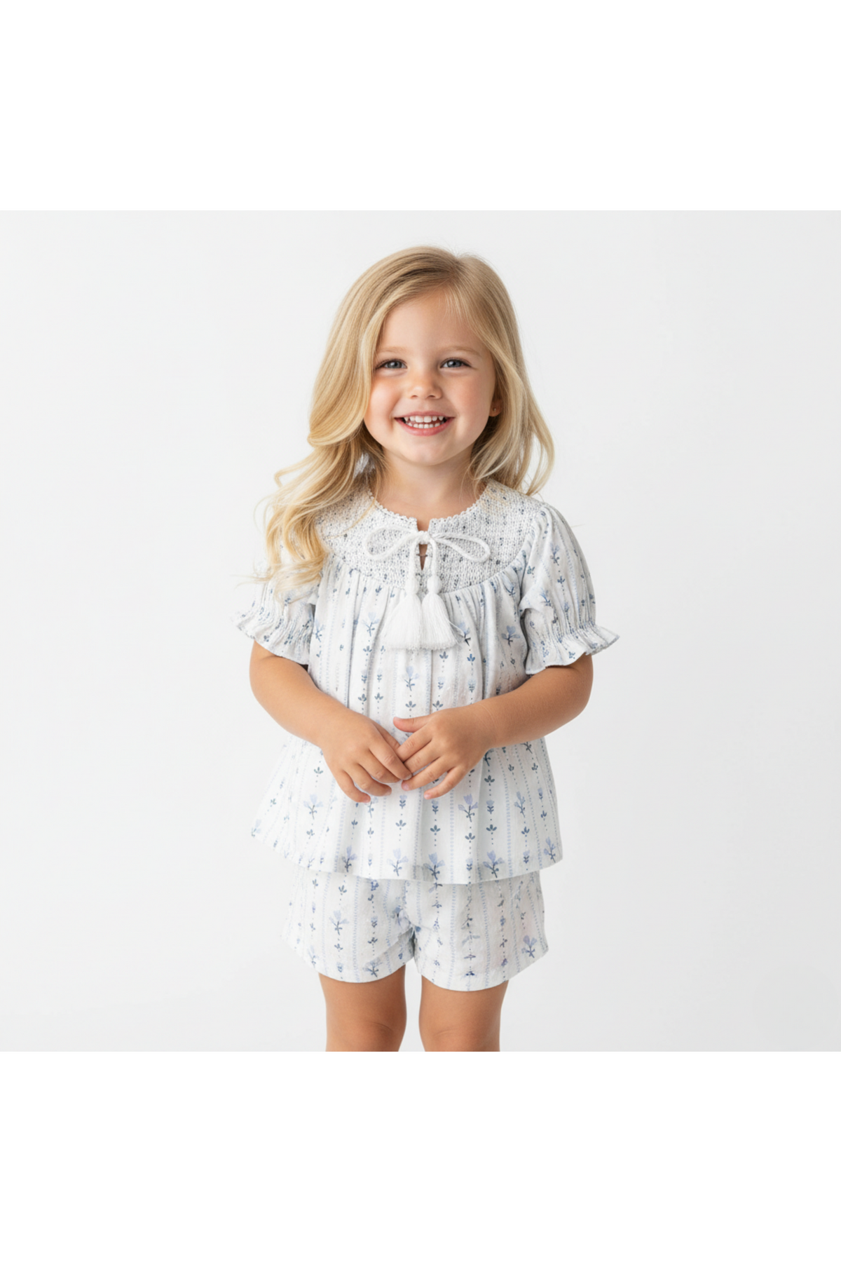 A smiling girl wearing a blue and white organic cotton smocked top with delicate embroidery and matching shorts, showcasing a playful and breezy style perfect for sunny day outings.