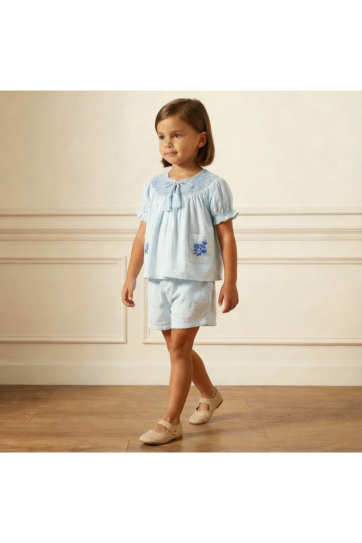 A young girl walks confidently in a light blue organic smocked top and matching shorts set, featuring delicate embroidery on the pocket, set against a softly lit, neutral background.