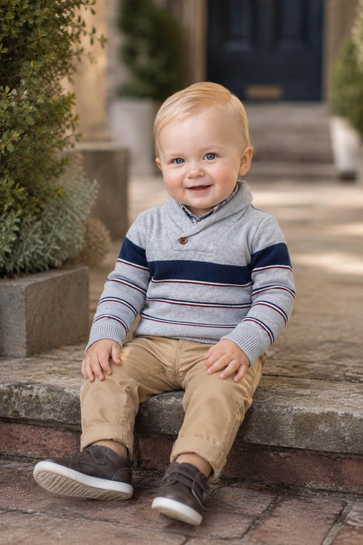 A smiling baby boy sits on stone steps, wearing a gray shawl collar sweater with navy and burgundy stripes, paired with beige pants and brown shoes.