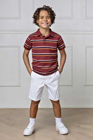 A young boy smiles while wearing a burgundy and striped polo shirt with white shorts, standing in a bright interior.