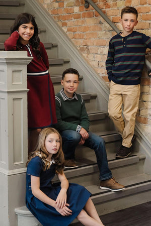 Four children pose on a staircase, wearing stylish outfits: a red dress, a green sweater, and casual pants.