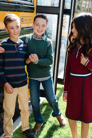 Three children stand outside a yellow school bus, smiling and interacting. One boy wears a green organic cotton sweater, while the others sport striped and maroon outfits, showcasing a stylish.