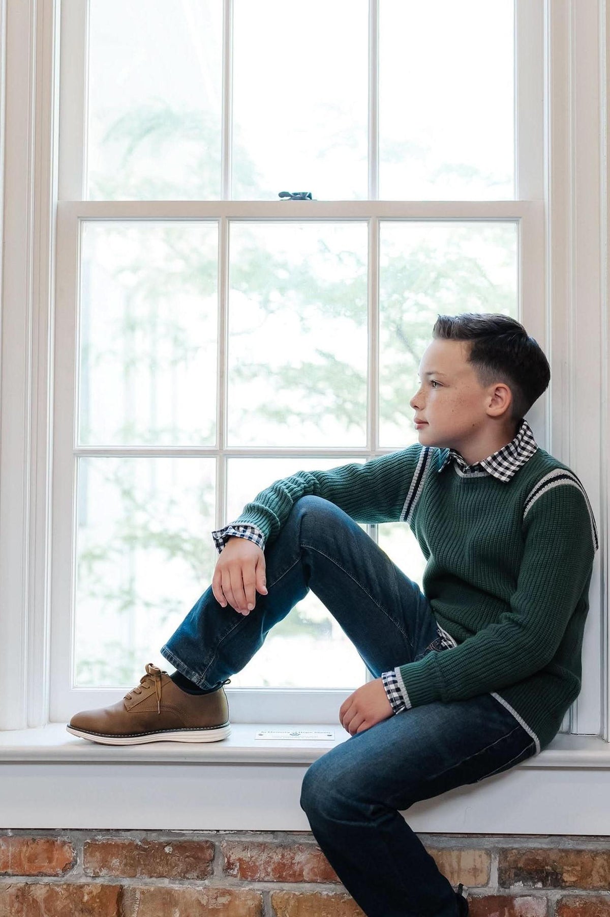 A young boy sits on a windowsill, wearing a green organic cotton sweater with sporty stripes and a checkered shirt underneath. He pairs it with blue jeans and brown shoes.