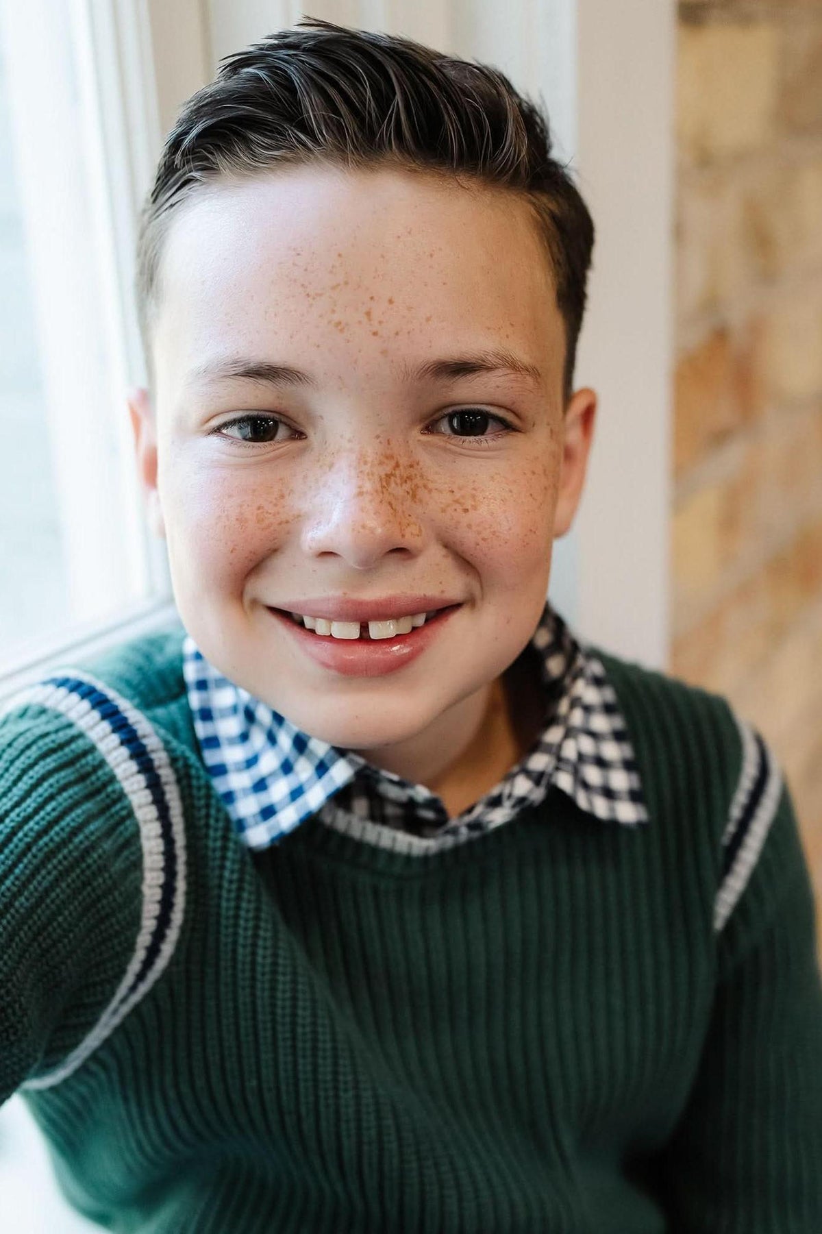 A smiling boy with freckles wears a green knit sweater over a navy gingham shirt, sitting by a window.