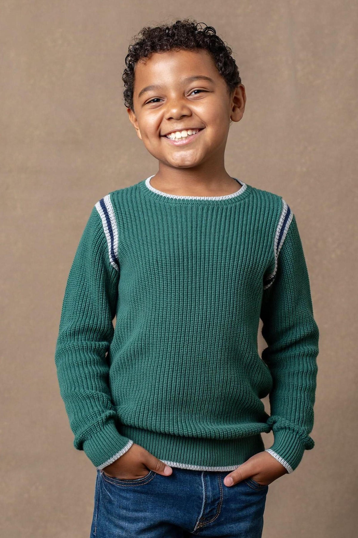 A smiling child wearing a green ribbed crew neck sweater with sporty stripe details, standing against a neutral background.