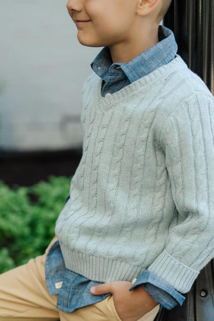 A young boy smiles while wearing a soft, dusty blue organic cotton cable knit V-neck sweater over a chambray shirt, paired with beige pants.